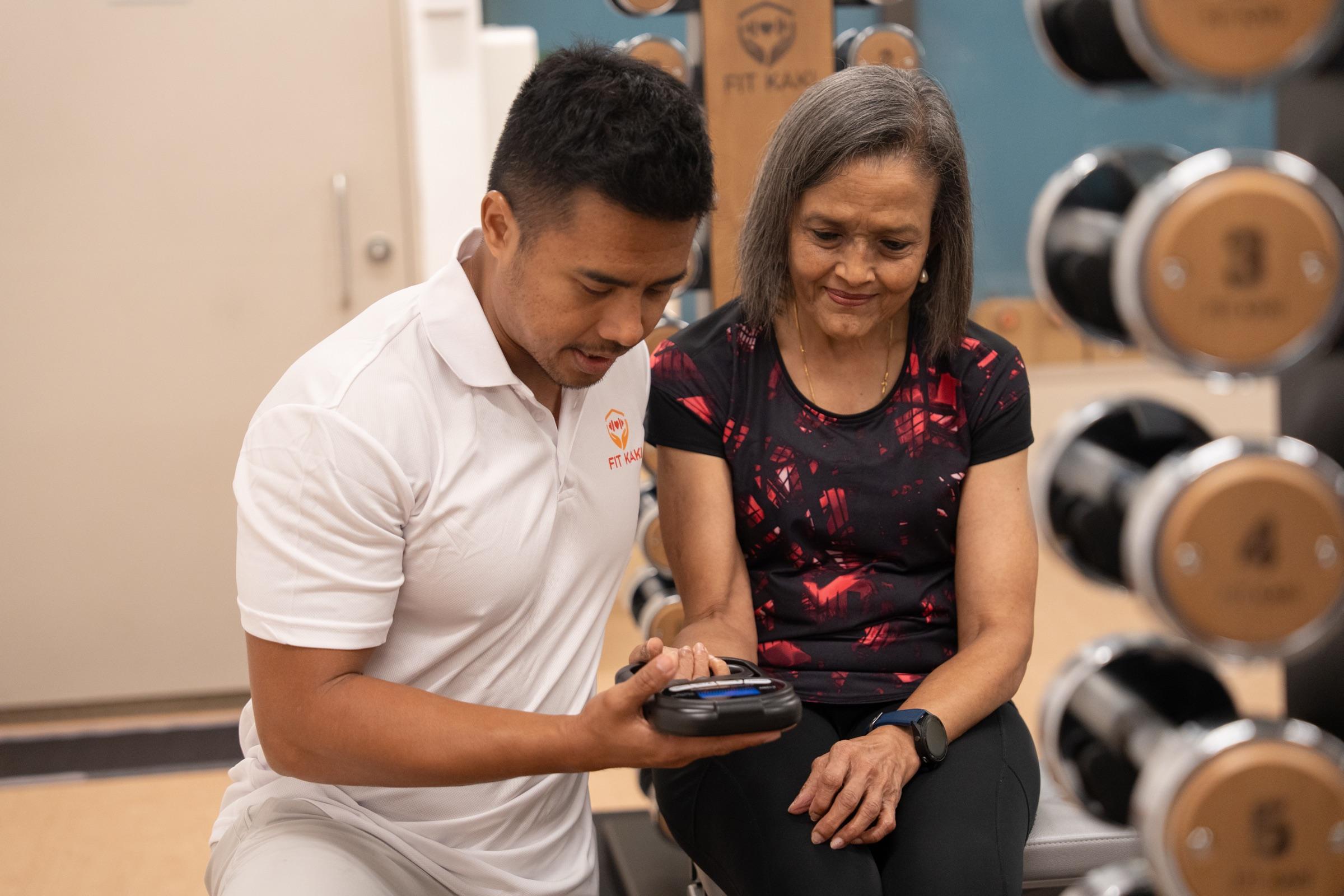 A Fit Kaki coach sitting beside a senior, patiently guiding her through a grip strength check at the Eunos studio